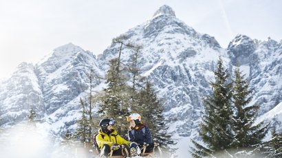 Paar sitzt mit Schlitten in verschneiter Berglandschaft vor schneebedeckten Gipfeln