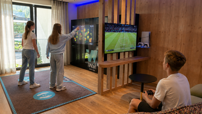 Zwei Mädchen spielen an einem interaktiven Bildschirm, Junge spielt Videospiel auf Fernseher