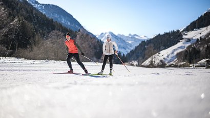Zwei Personen beim Skilanglauf vor verschneiten Bergen bei sonnigem Wetter