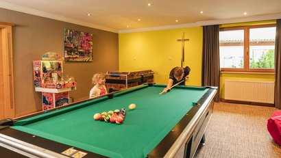Kinder spielen Billard in einem hellen Spielzimmer mit Fenster und Spielautomat