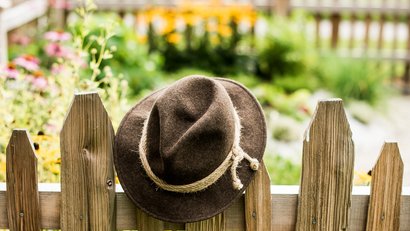 Brauner Filzhut hängt an einem Holzzaun vor einem Blumenbeet