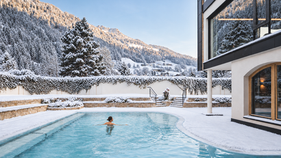 Person schwimmt im Außenpool eines Hotels, umgeben von Schnee und verschneiten Bergen