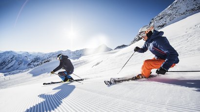 Zwei Skifahrer fahren bei Sonnenschein auf frisch präparierter Piste in den Bergen