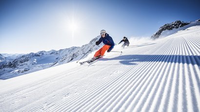 Zwei Skifahrer fahren auf frisch gespurter Piste in sonniger Berglandschaft