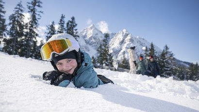 Kind im Schnee liegend mit Skibrille neben verschneiter Berglandschaft
