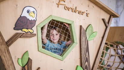 Kind schaut durch ein Netzfenster in einem hölzernen Spielhaus mit Adlerbild