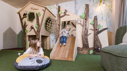 Kinder spielen auf einer Holzrutsche in einem spielerischen Raum mit Waldmotiven